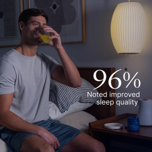 Man sitting on a bed in a dimly lit room with a lamp and nightstand, drinking from a glass.