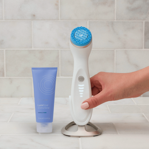White facial cleansing brush lumispa with blue bristle head on a tiled bathroom floor next to a tube of Lumispa Skin cleanser.