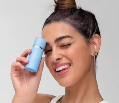 Woman holding a blue blemish serum product against her face on a plain background