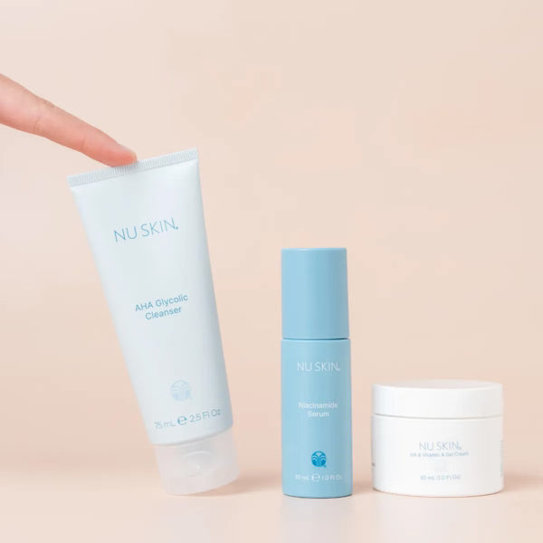 Three Nu Skin skincare products arranged on a pale peach background: a finger points to the large light blue AHA Glycolic Cleanser tube, next to a light blue Absorbing Serum bottle and a white cream jar.