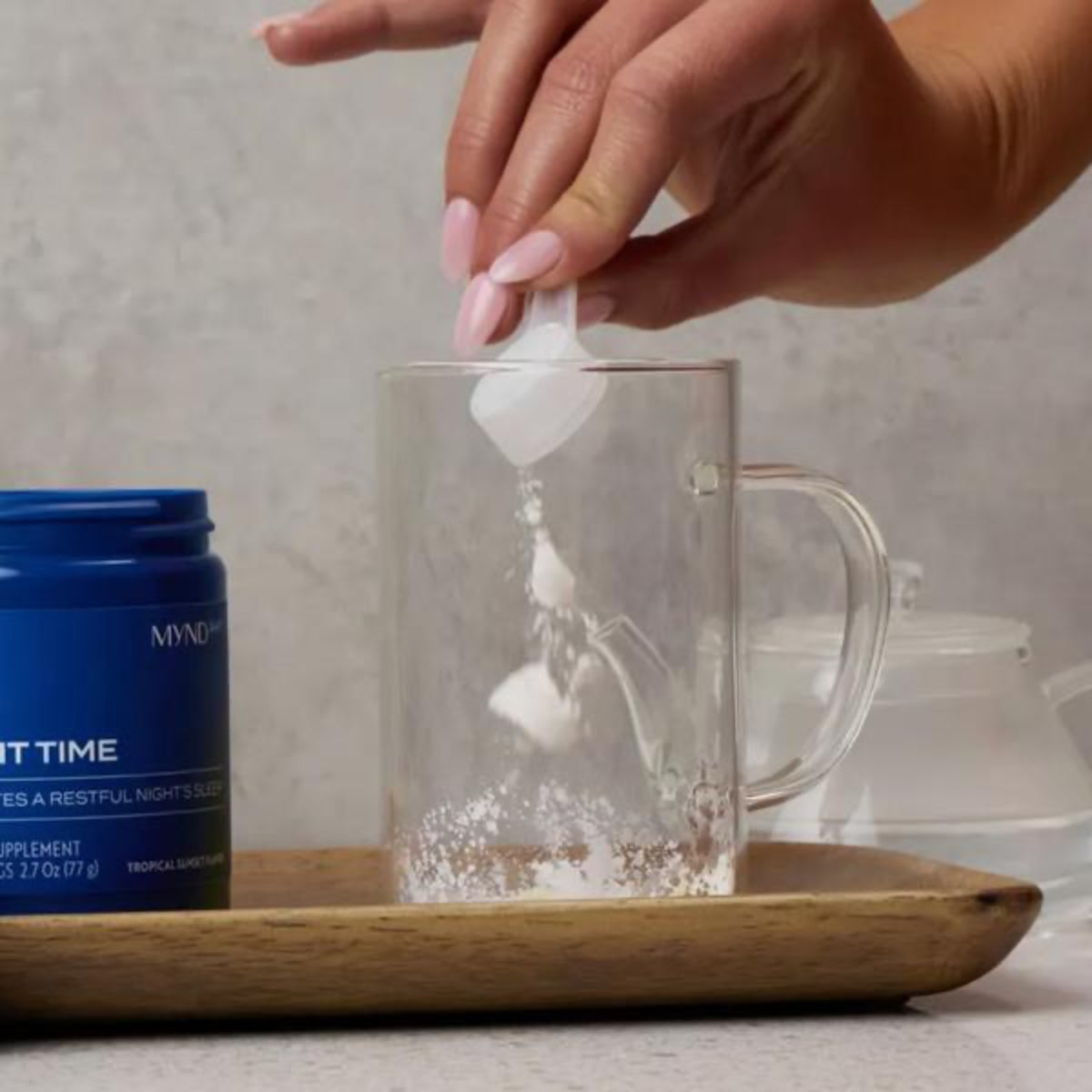 Hand adding a tea bag into a clear mug with a Myndful supplement container on a wooden tray.