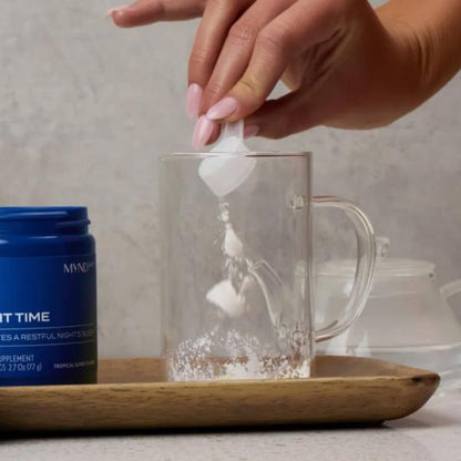 Hand adding a tea bag into a clear mug with a Myndful supplement container on a wooden tray.